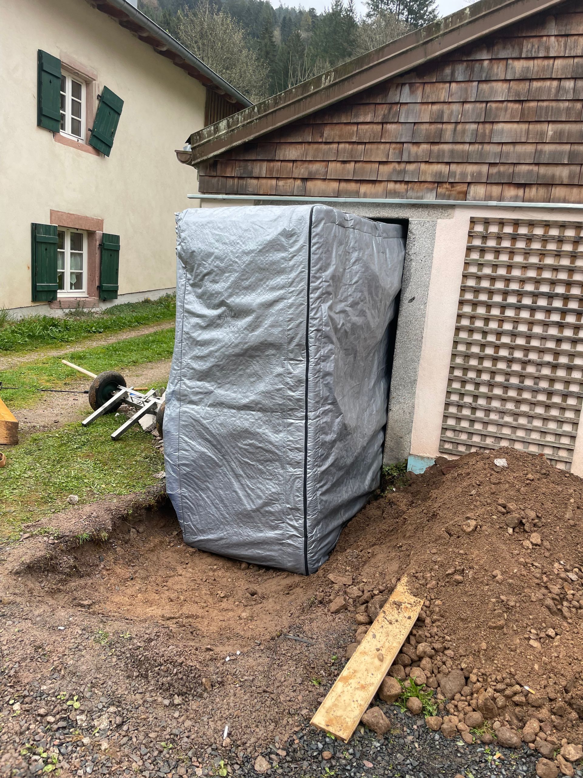 Un hangar est recouvert d'une bâche et posé dans la terre devant une maison.