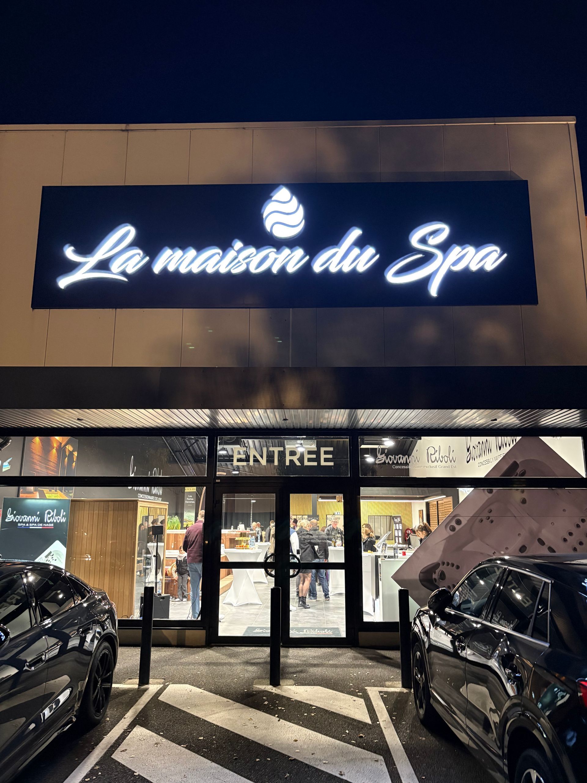 L'enseigne « La Maison du Spa » est illuminée la nuit. Le panneau « Entrée » est au-dessus de la porte. Voitures garées à proximité.