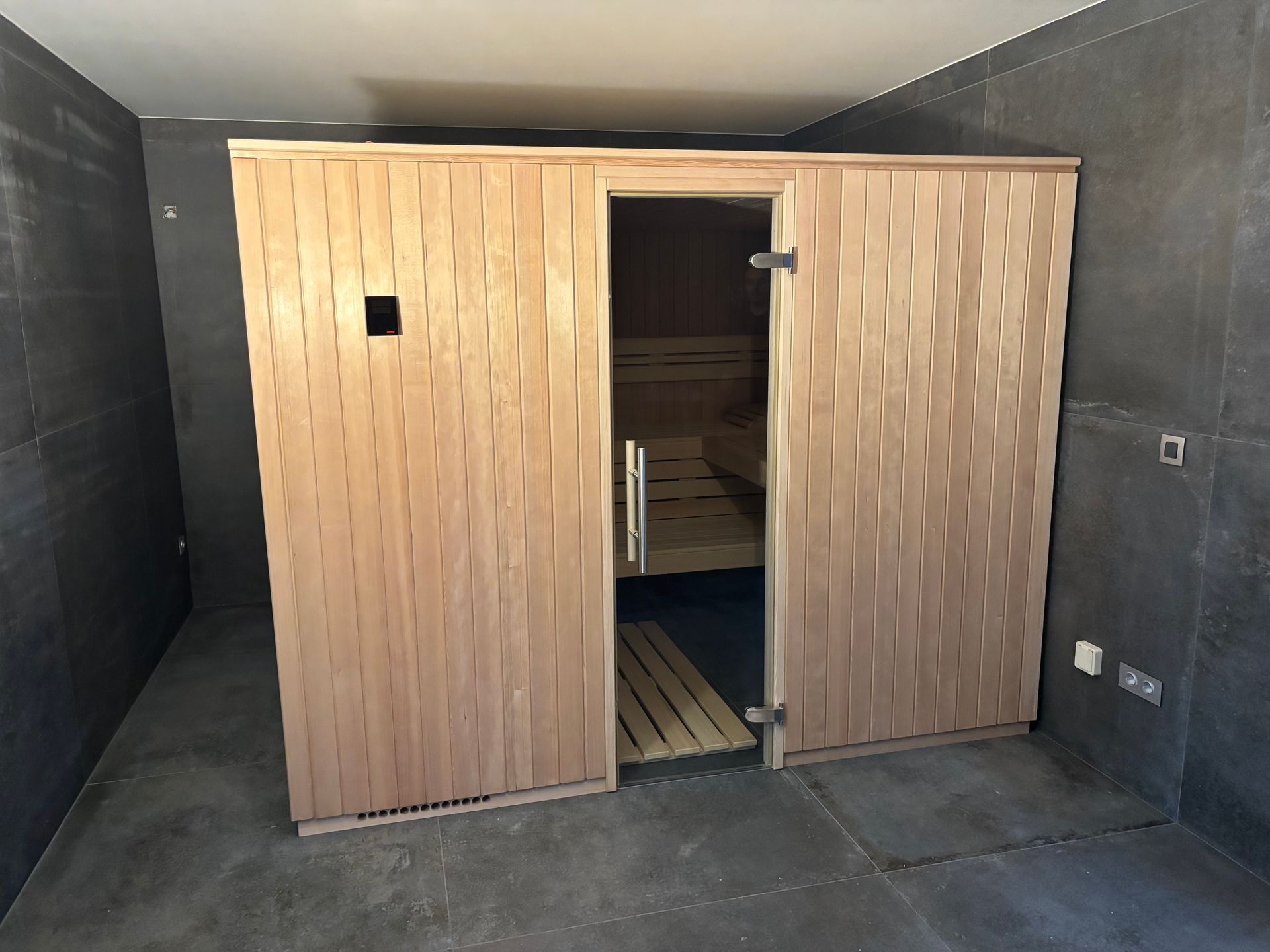 Sauna en bois avec porte ouverte dans une pièce aux murs et au sol gris foncé.