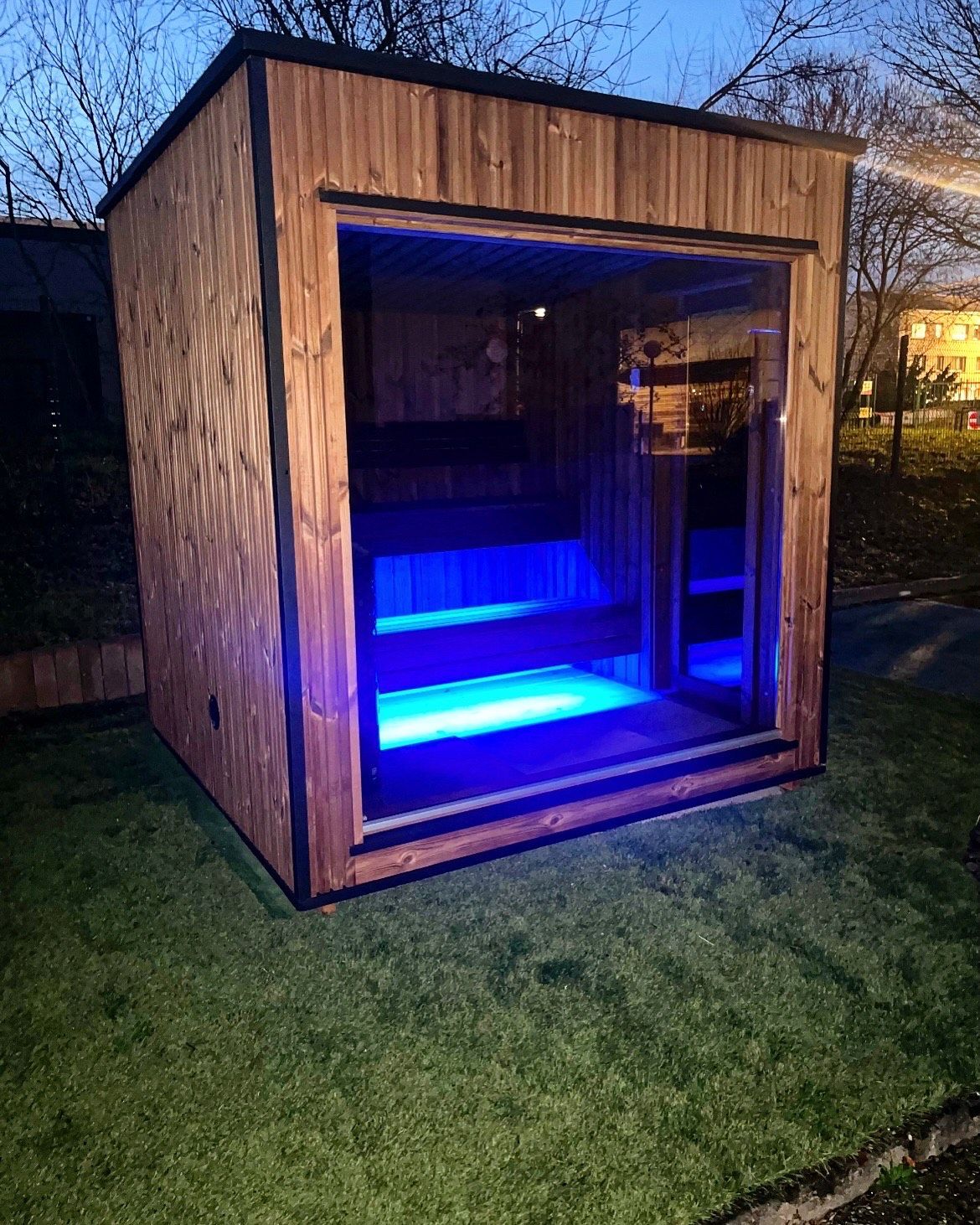 Un hangar en bois avec des lumières bleues dessus est posé sur une pelouse verte luxuriante.