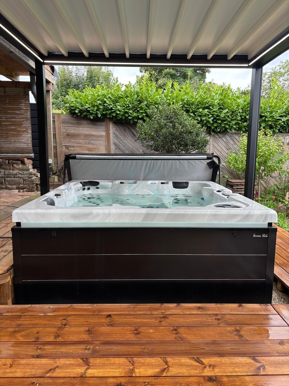 Jacuzzi sous une pergola sur une terrasse en bois. Le jacuzzi est marron foncé, équipé de jets et d'un couvercle partiellement ouvert.
