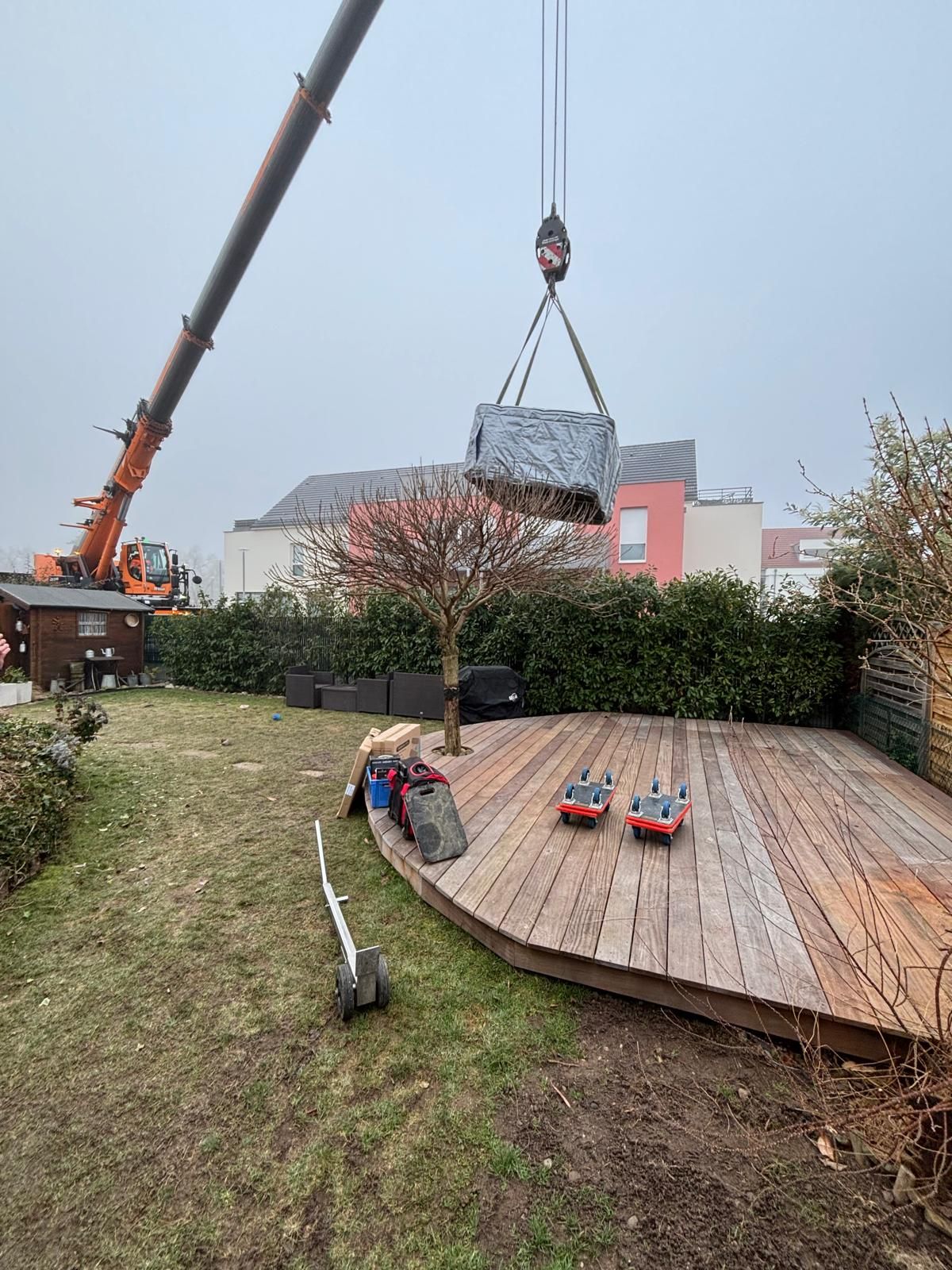 Une grue soulevant un grand objet emballé au-dessus d'une terrasse arrière avec des bâtiments en arrière-plan.