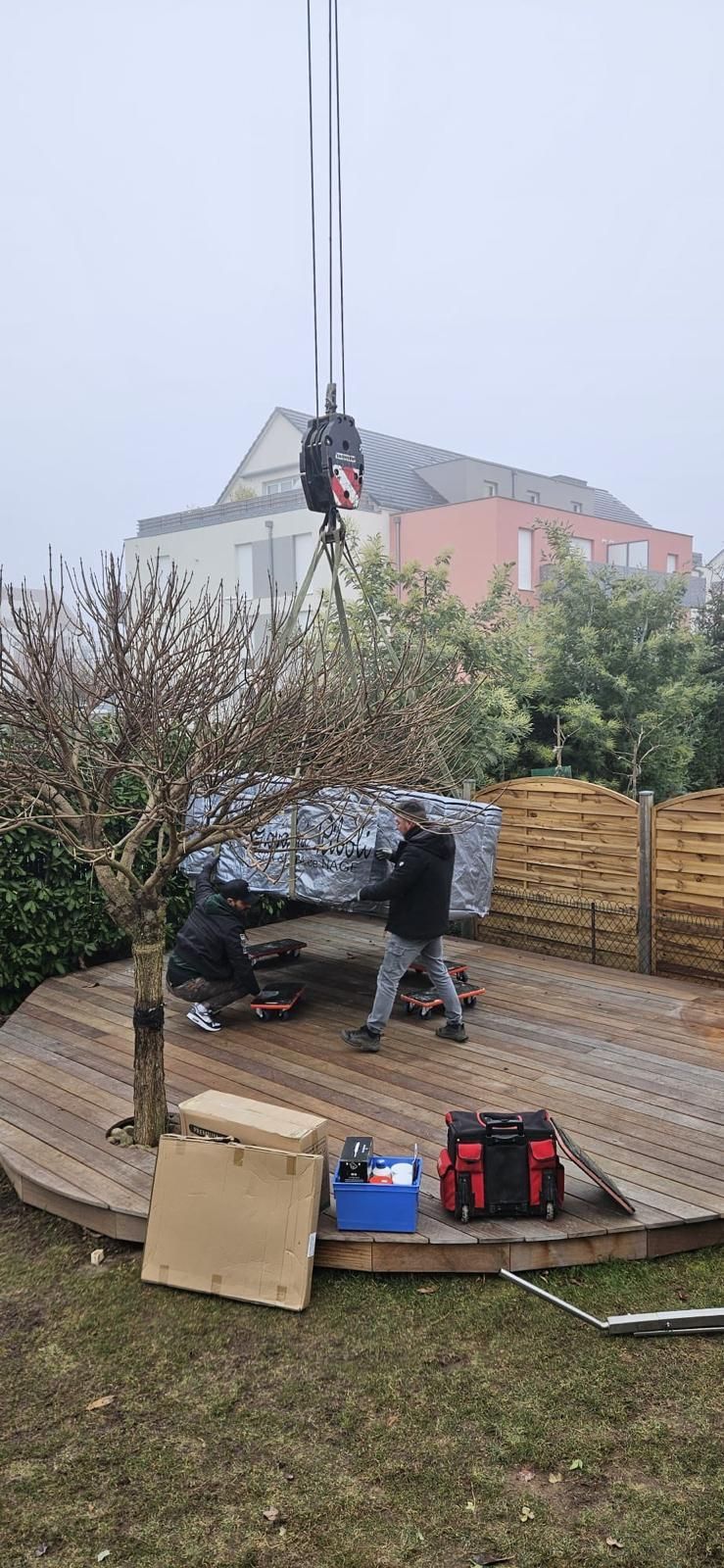 Une grue soulève un objet couvert sur une terrasse en bois dans un jardin. Deux personnes sont présentes, avec des maisons en arrière-plan.