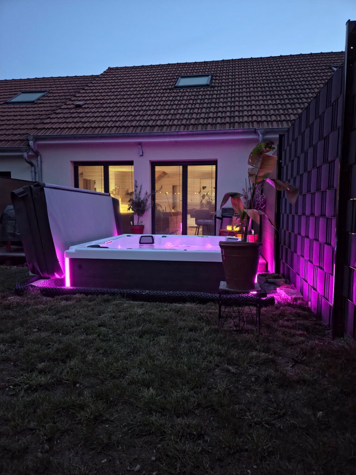 Jacuzzi avec lumières violettes, installé dans un jardin au crépuscule. Maison avec fenêtres éclairées en arrière-plan.