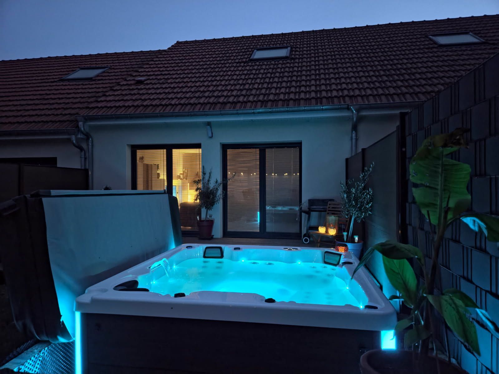 Un bain à remous éclairé brille en bleu au crépuscule, placé devant une maison avec des portes coulissantes en verre ouvertes et un toit en tuiles.