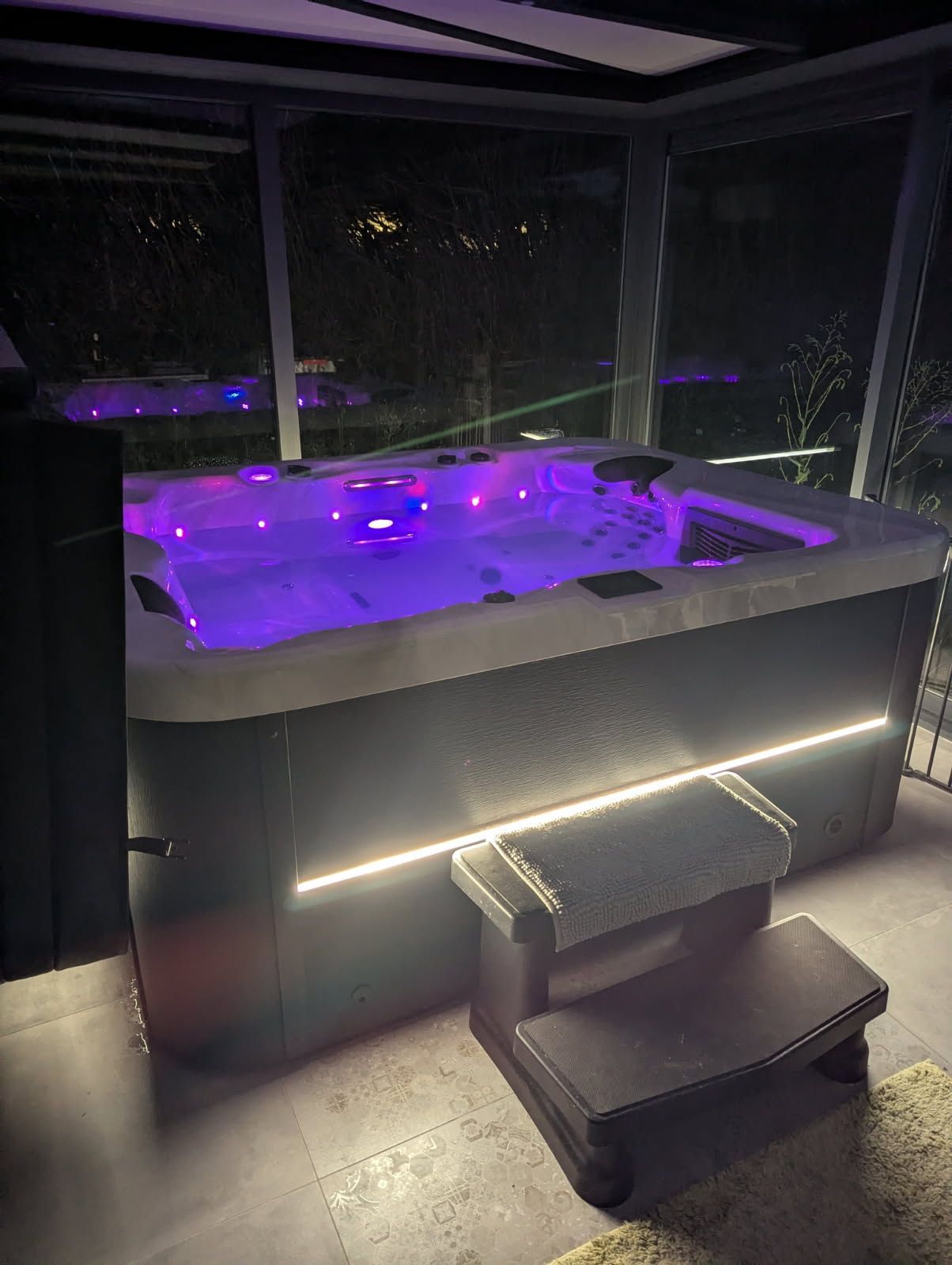 Spa éclairé avec des lumières violettes et un marchepied, à l'extérieur la nuit.