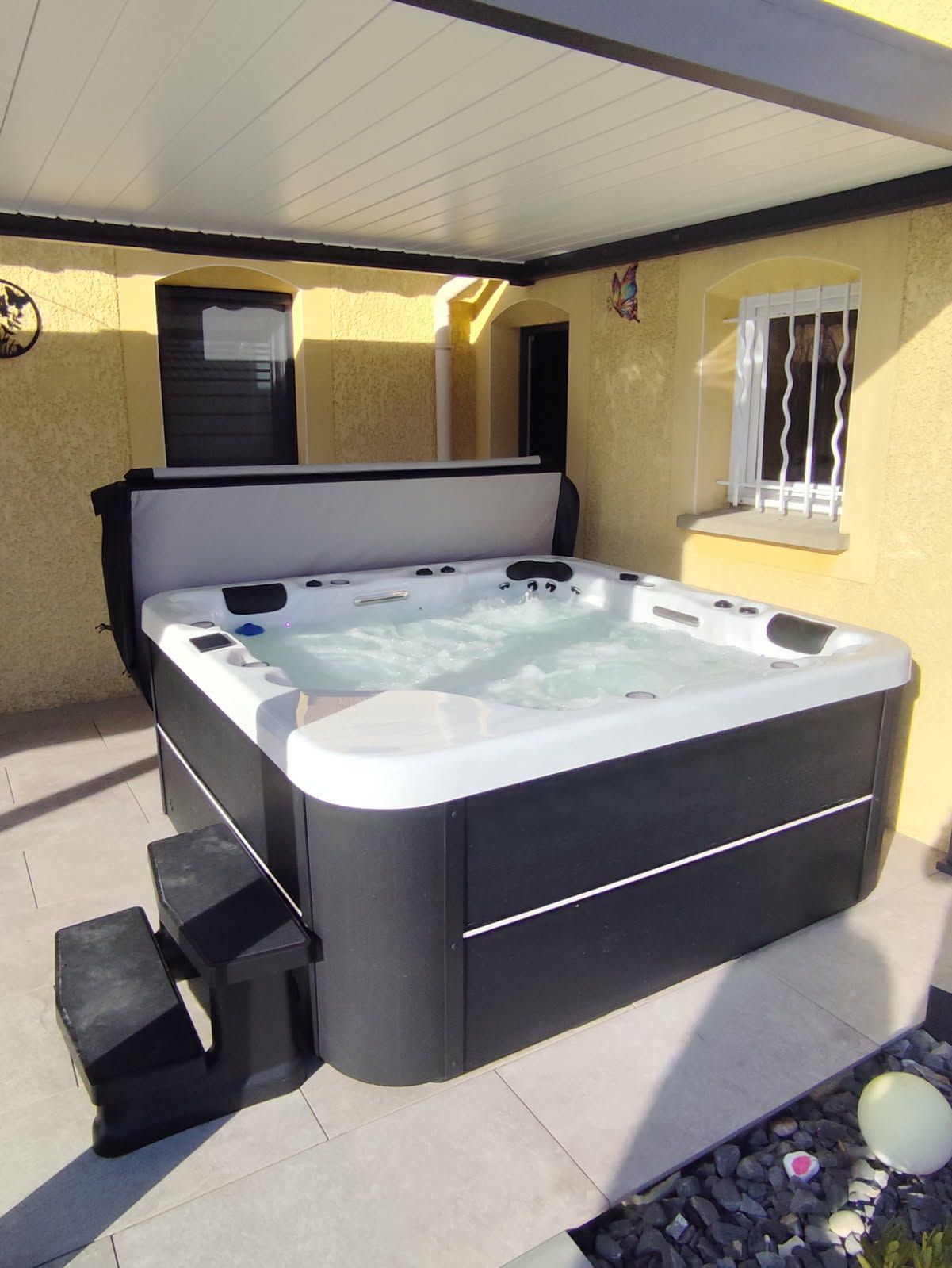 Jacuzzi sous une terrasse couverte, avec marches devant. Jacuzzi noir et blanc sur fond de bâtiment jaune et de terrasse grise.