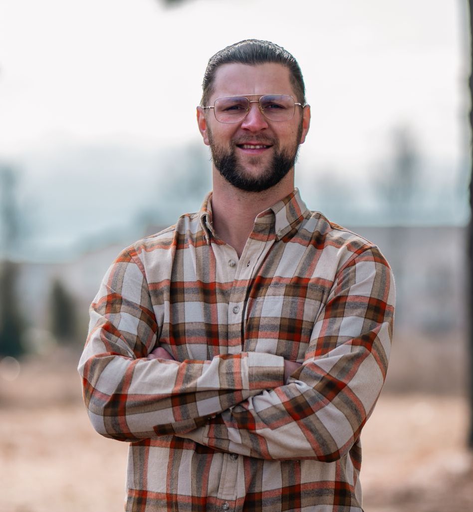 A man with a beard wearing glasses and a plaid shirt is standing with his arms crossed.