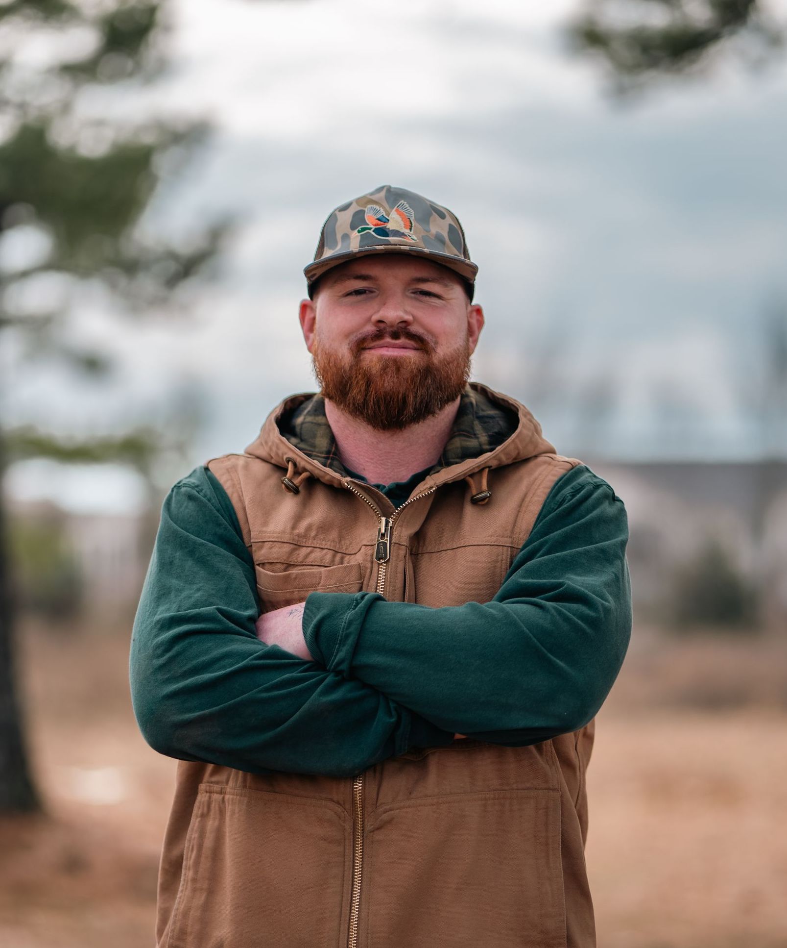 A man with a beard and a hat is standing with his arms crossed.