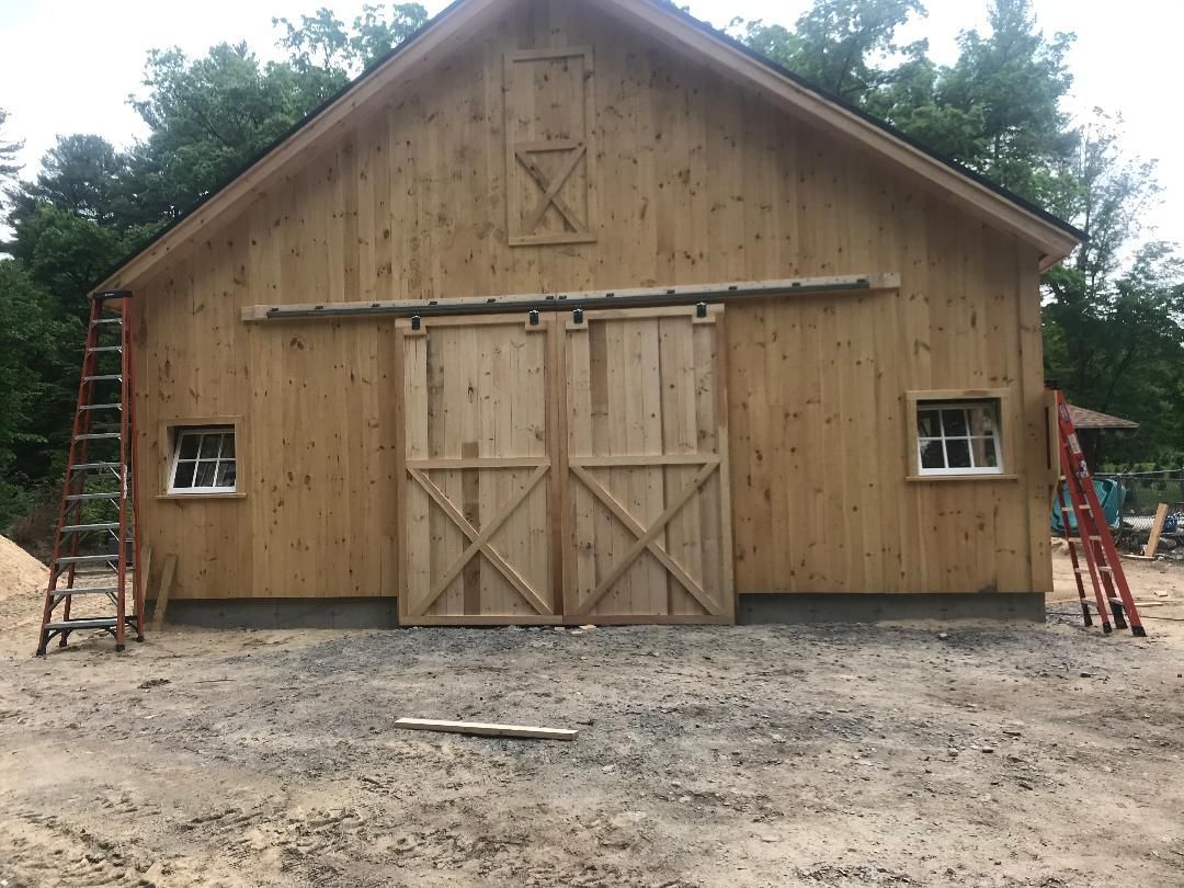 Wooden barn with sliding doors, two small windows, and ladders on both sides, built on a gravel surface.