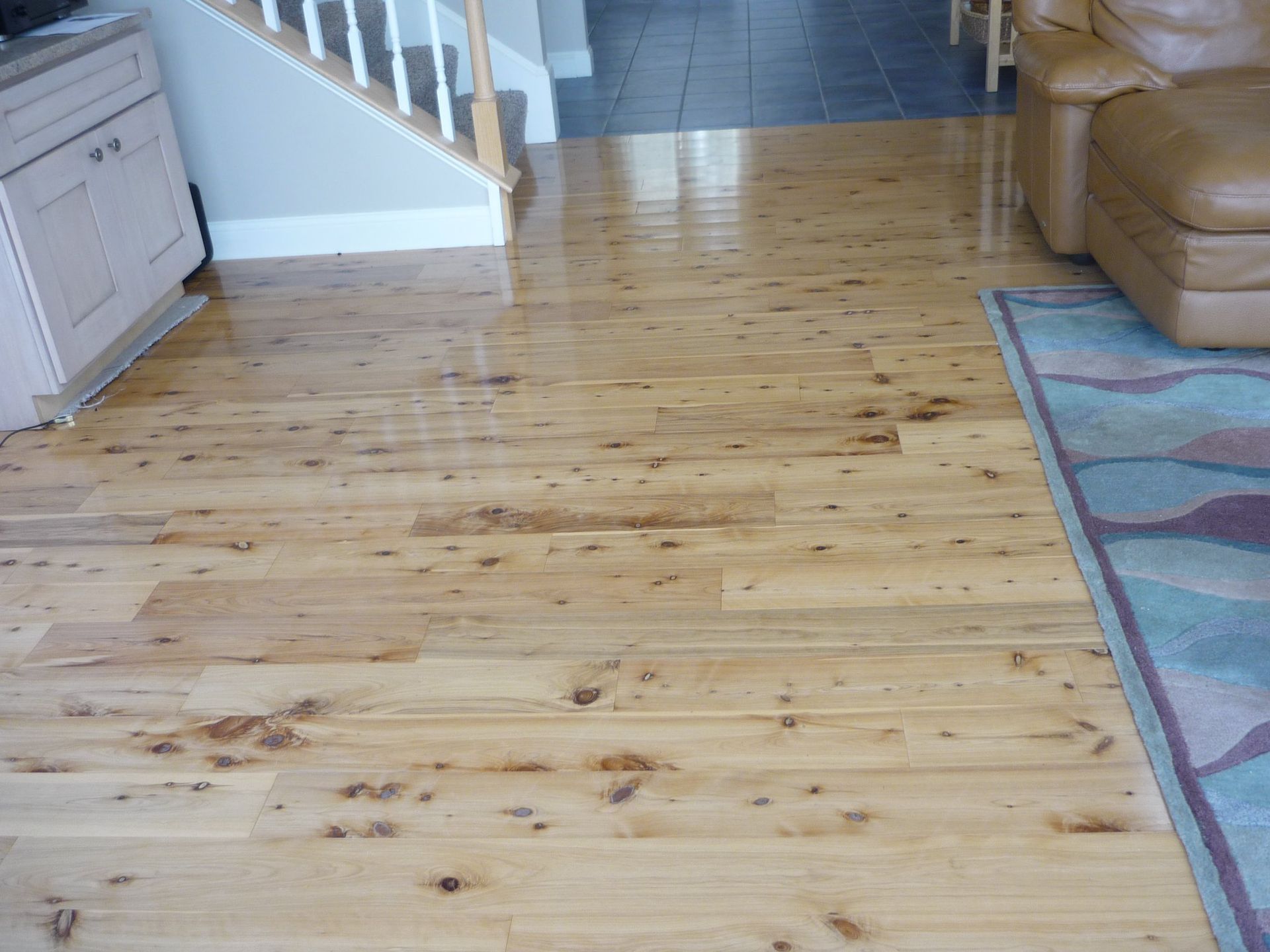 Light wood floor in a home, reflecting light. A rug and leather couch are visible.