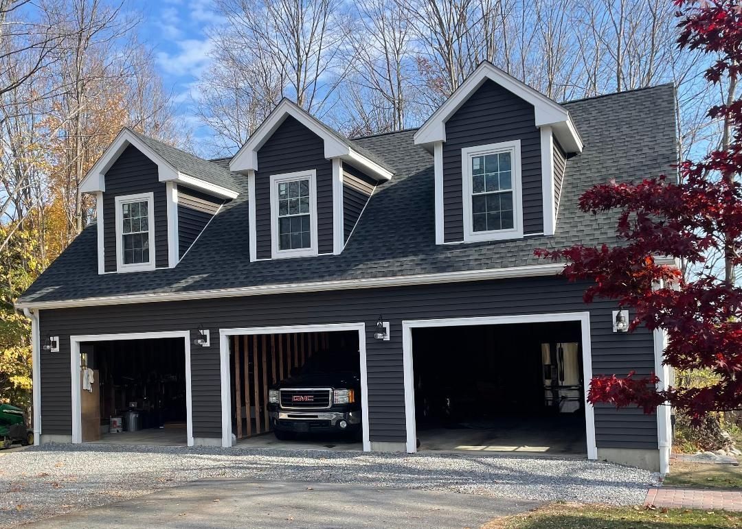 Dark gray three-car garage with three dormers, white trim, and a gravel driveway. A truck is parked inside.