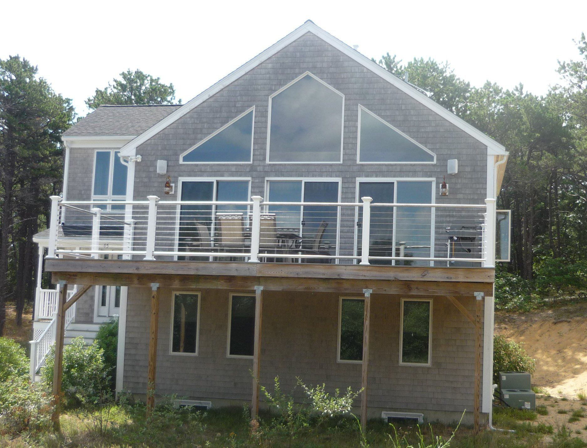 Two-story gray shingled house with deck overlooking a forest. Large windows, glass railing.
