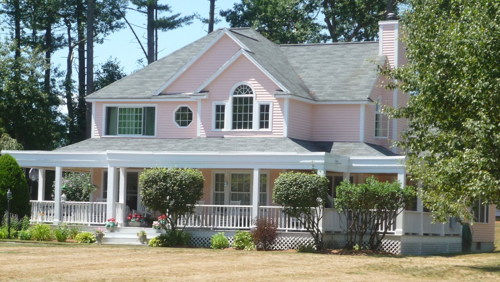 Pink two-story house with a wraparound porch, set on a large lawn, with trees in the background.