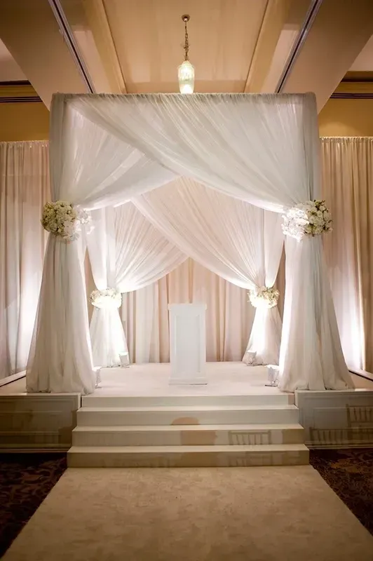 Elegant white wedding chuppah with draped fabric and floral accents on a raised platform.