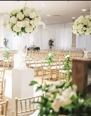 Wedding ceremony setup, white flowers, gold chairs, white drapery backdrop.