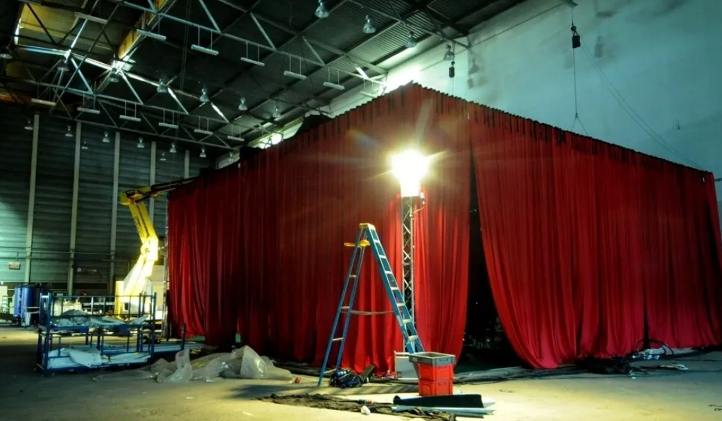 A large red curtain structure inside a dimly lit warehouse with a ladder and spotlight.