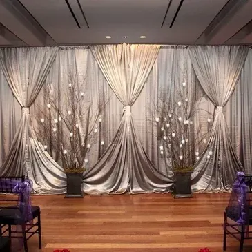 Wedding ceremony backdrop with silver draping, floral arrangements, and wooden floor.