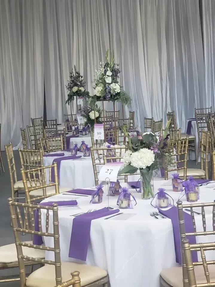 Wedding reception tables set with white tablecloths, purple runners, and gold chairs.