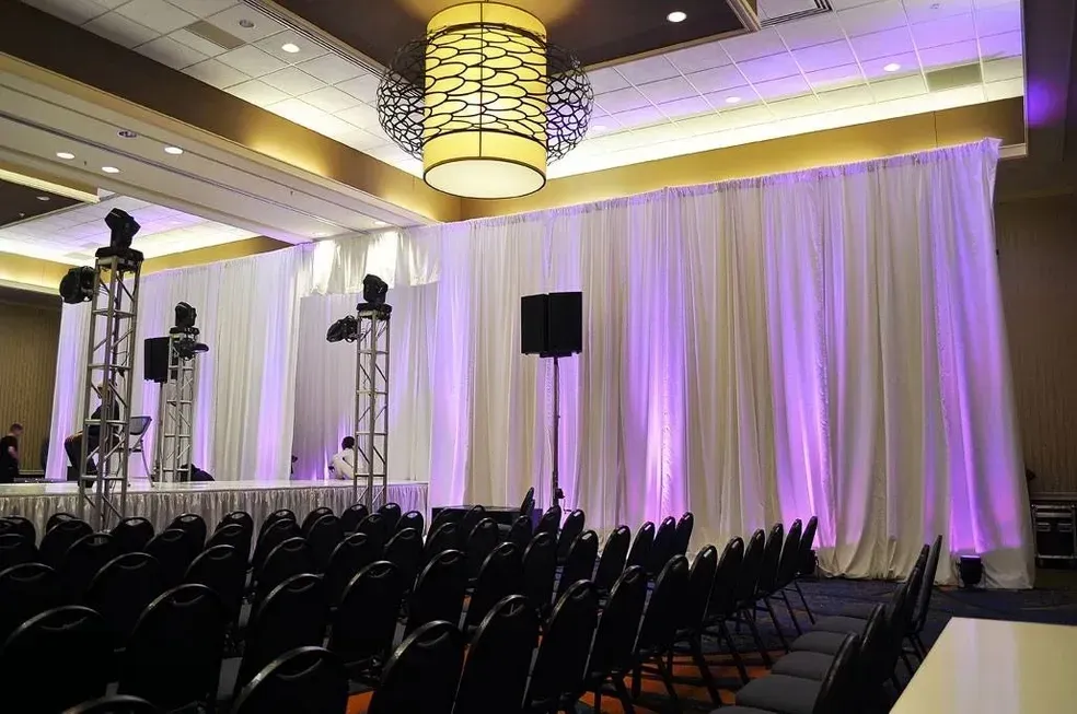 Conference stage with white draped backdrop, purple lighting, speakers, and rows of black chairs.