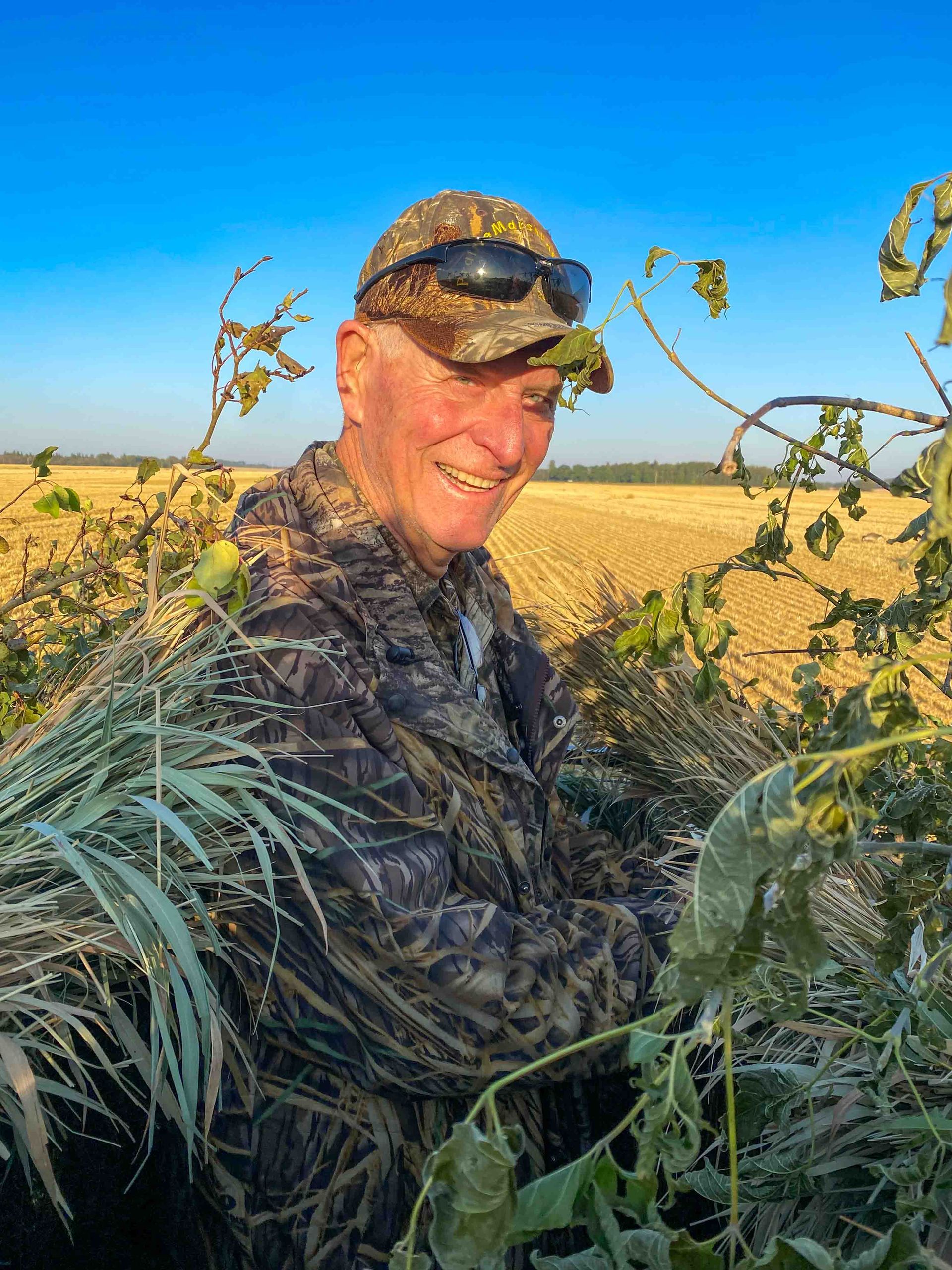 A man in a camouflage jacket and hat is standing in a field.