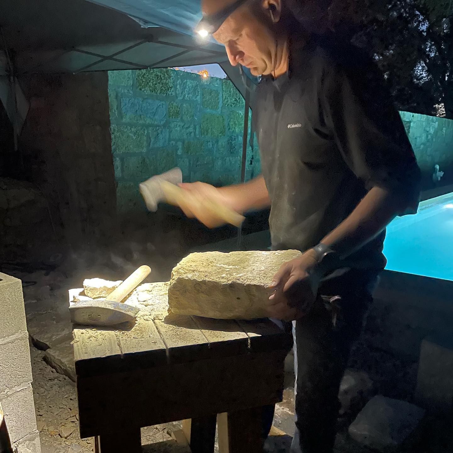 Man carving stone at night with hammer and chisel, lit by headlamp.