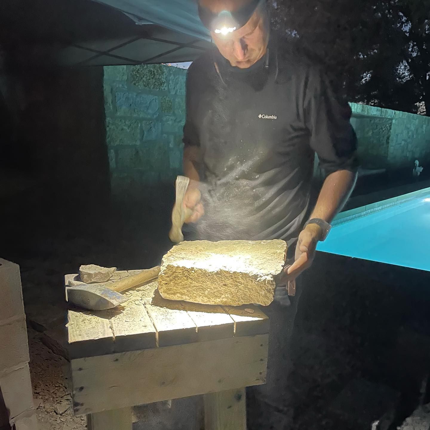 Man using a mallet and chisel on a brick outdoors at night, illuminated by a headlamp.