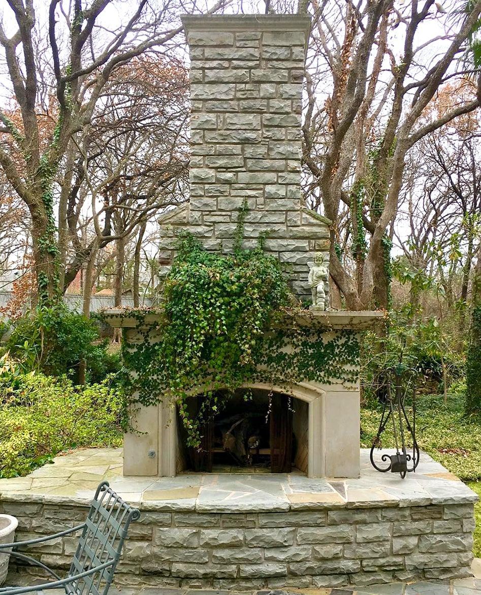 Outdoor stone fireplace with green vines, in a backyard with trees.