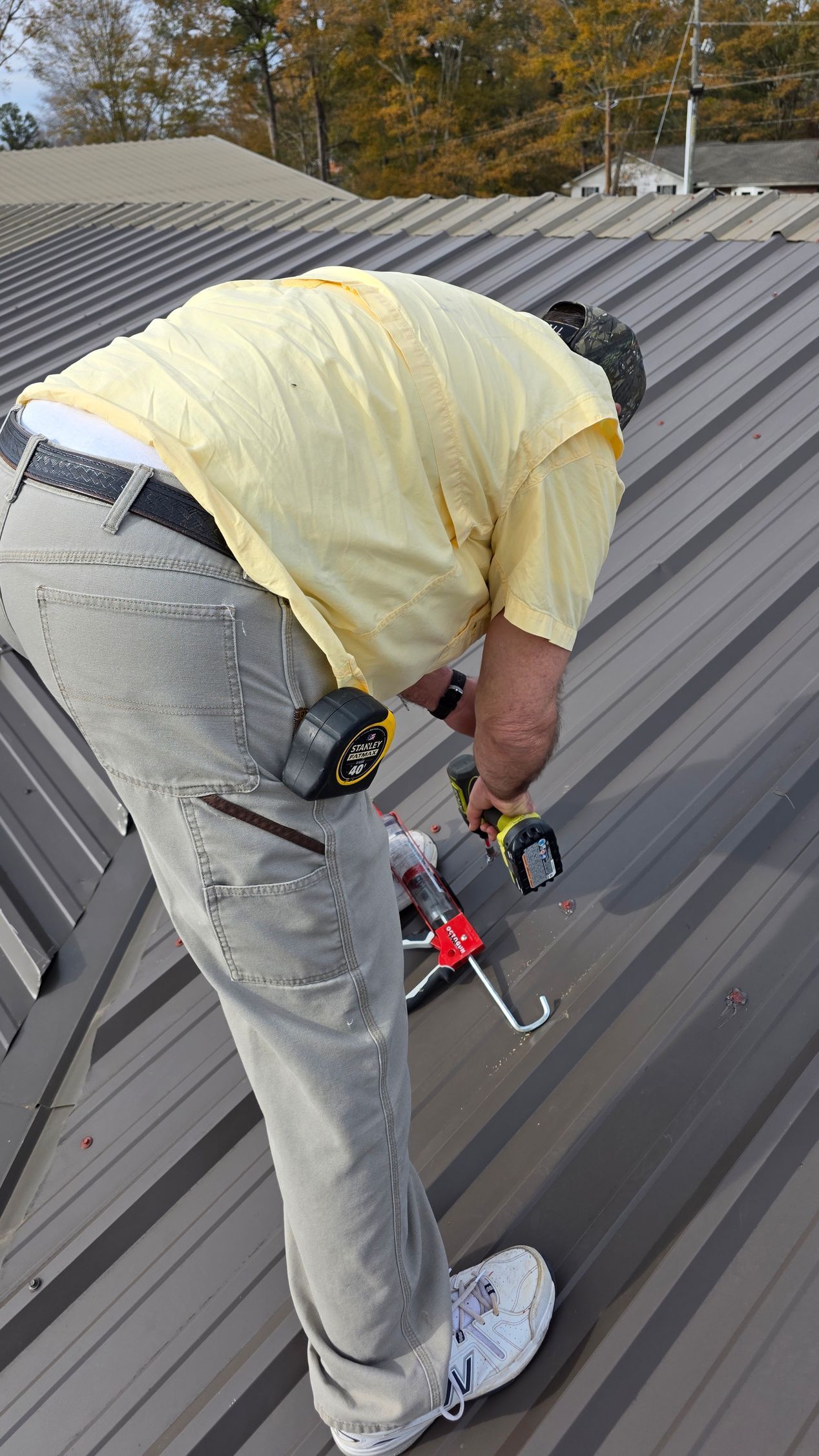 Worker on a metal roof using a power tool, wearing a yellow shirt and gray pants