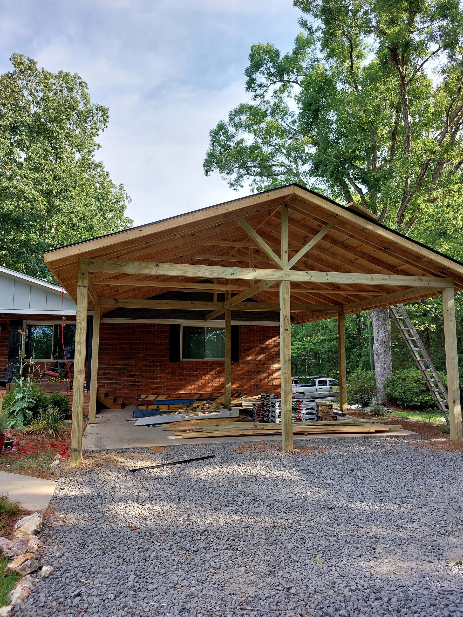Rustic wooden carport with gravel driveway, trees, and a small brick building in the background