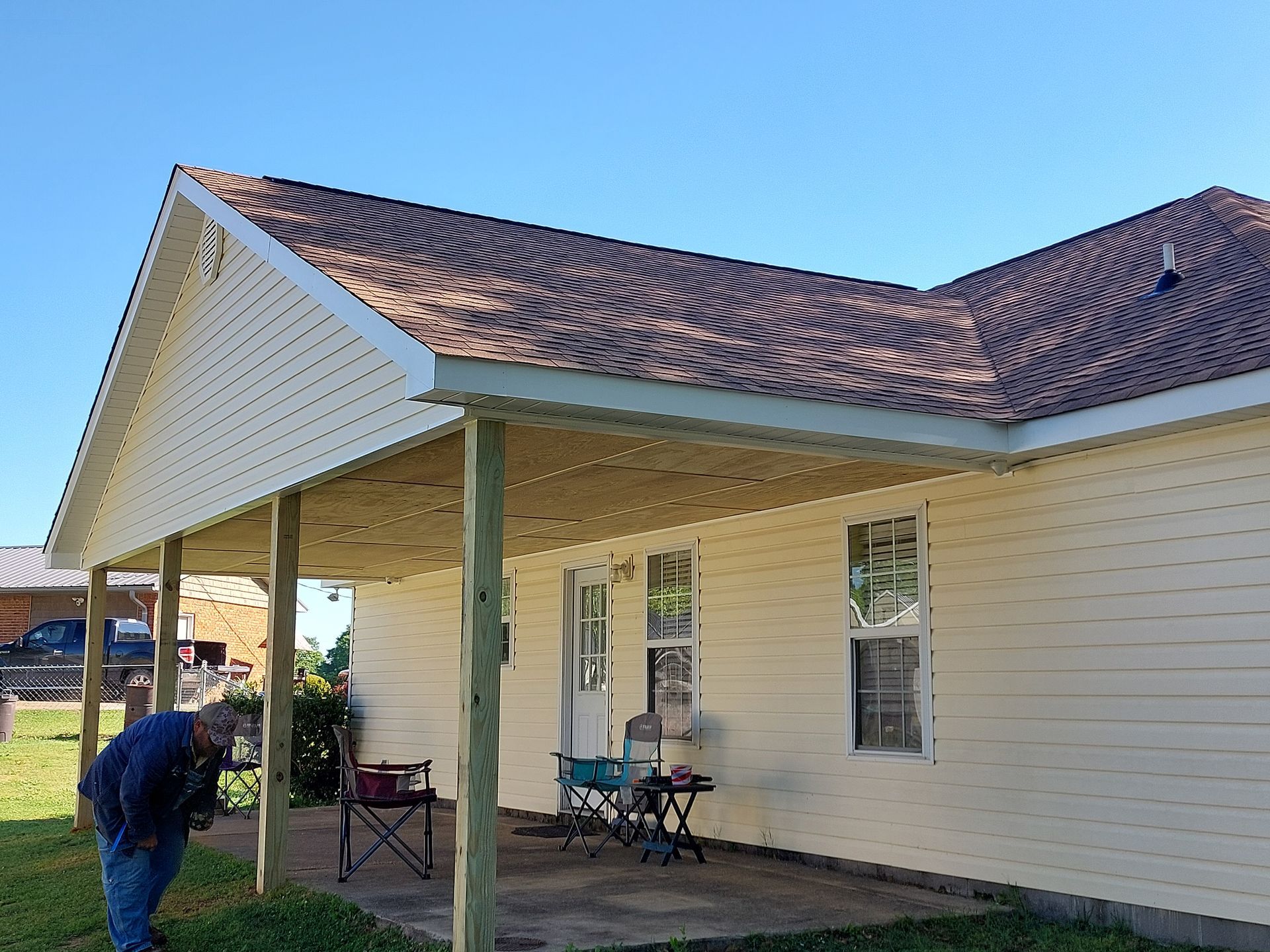 Porch of a cream house with a tiled roof; a person bends near a green post beside patio chairs.