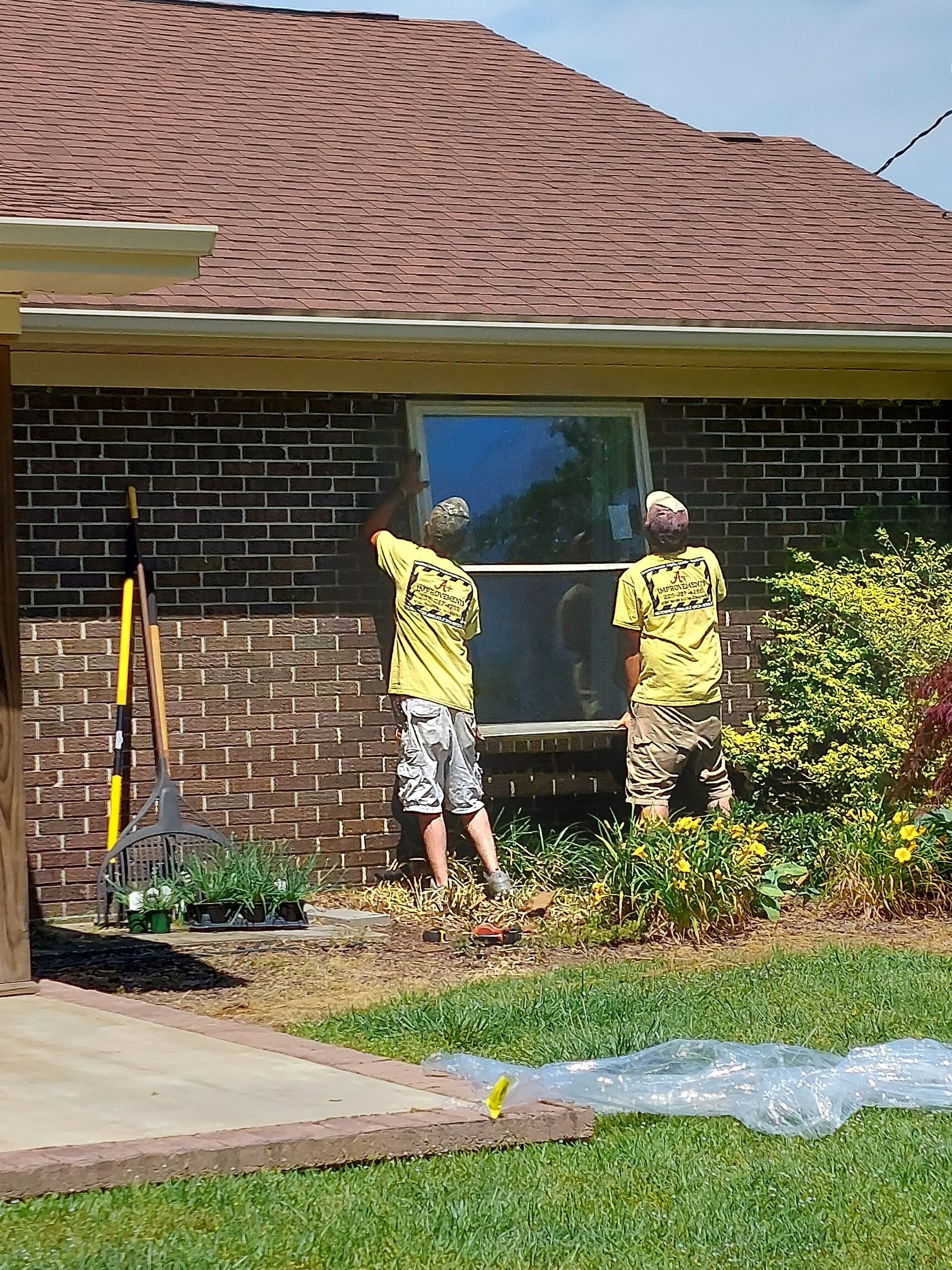Two workers in yellow shirts installing a window on a brick house exterior