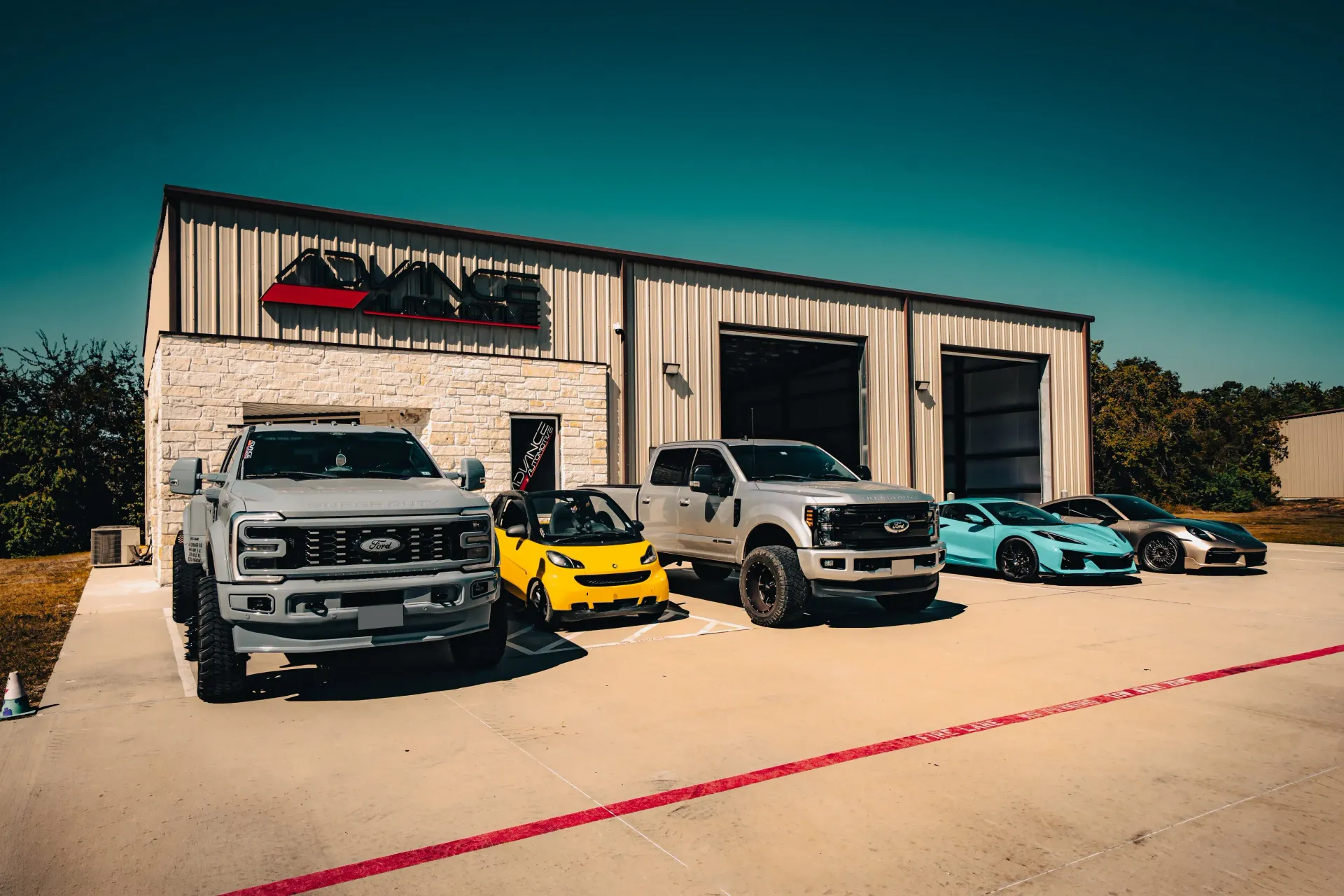 Vehicles parked outside a grey building with open bay doors. Cars include trucks and sports cars, on a sunny day. | Advance Automotive