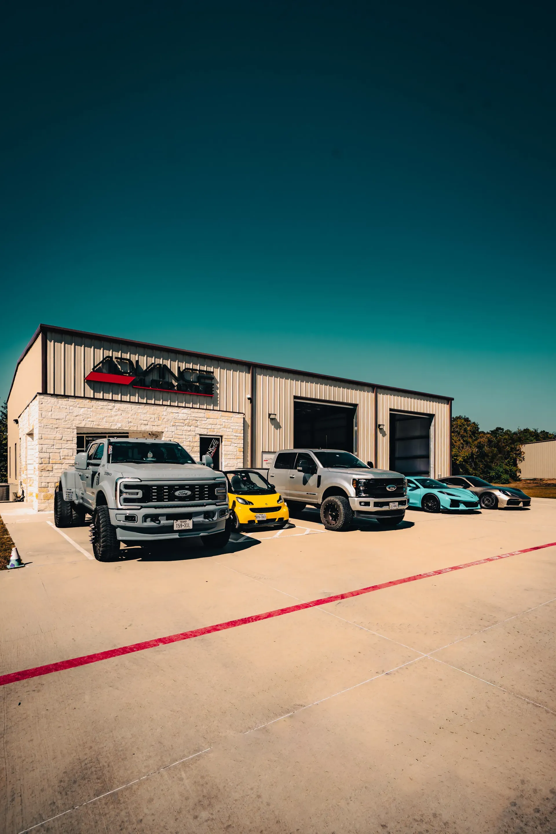 A building with a sign, several trucks, a yellow car, and sports cars parked outside on a sunny day. | Advance Automotive