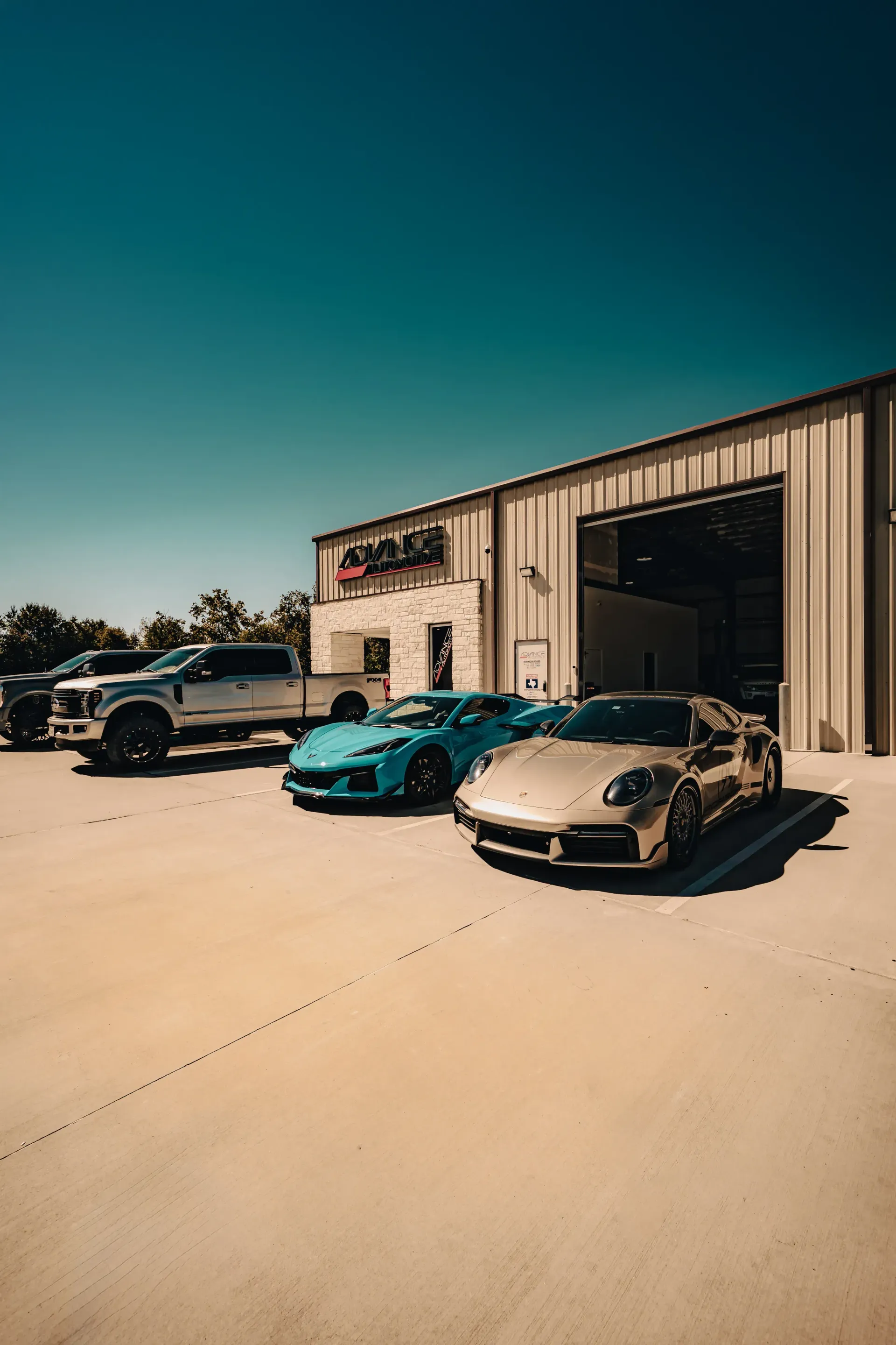 A light-colored sports car parked in front of a garage with a blue sports car and a pickup truck on a sunny day. | Advance Automotive