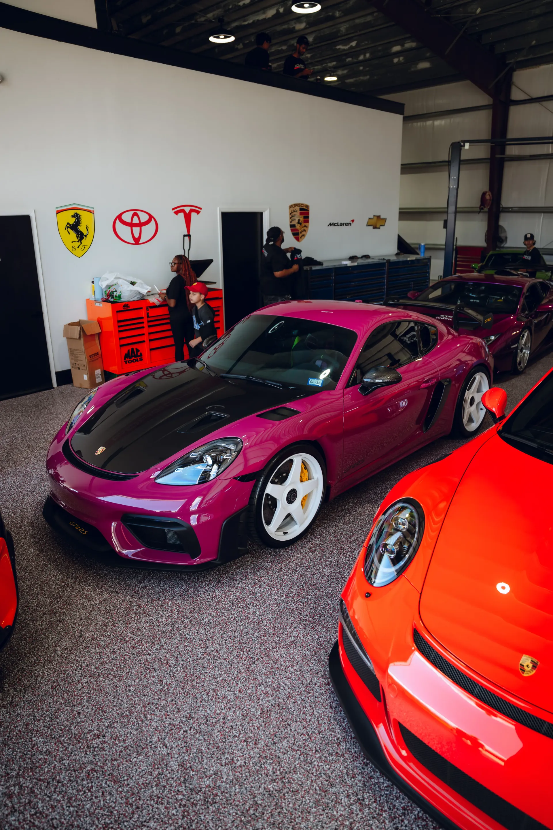A vibrant purple Porsche Cayman with a black hood sits in a garage with other sports cars. | Advance Automotive