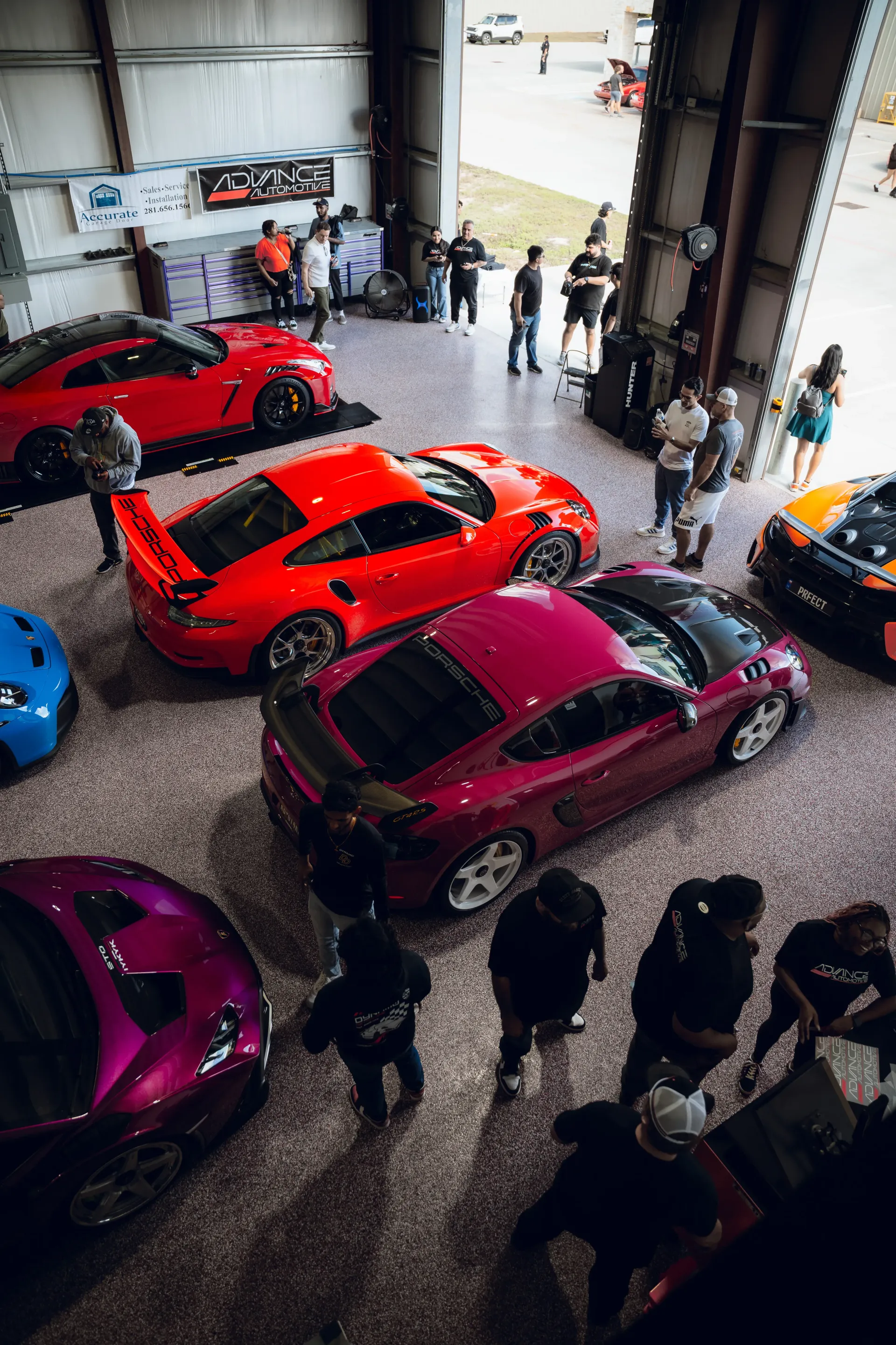 Cars on display at an event, with people. Various colors: red, pink, blue, orange. Inside a large open building. | Advance Automotive