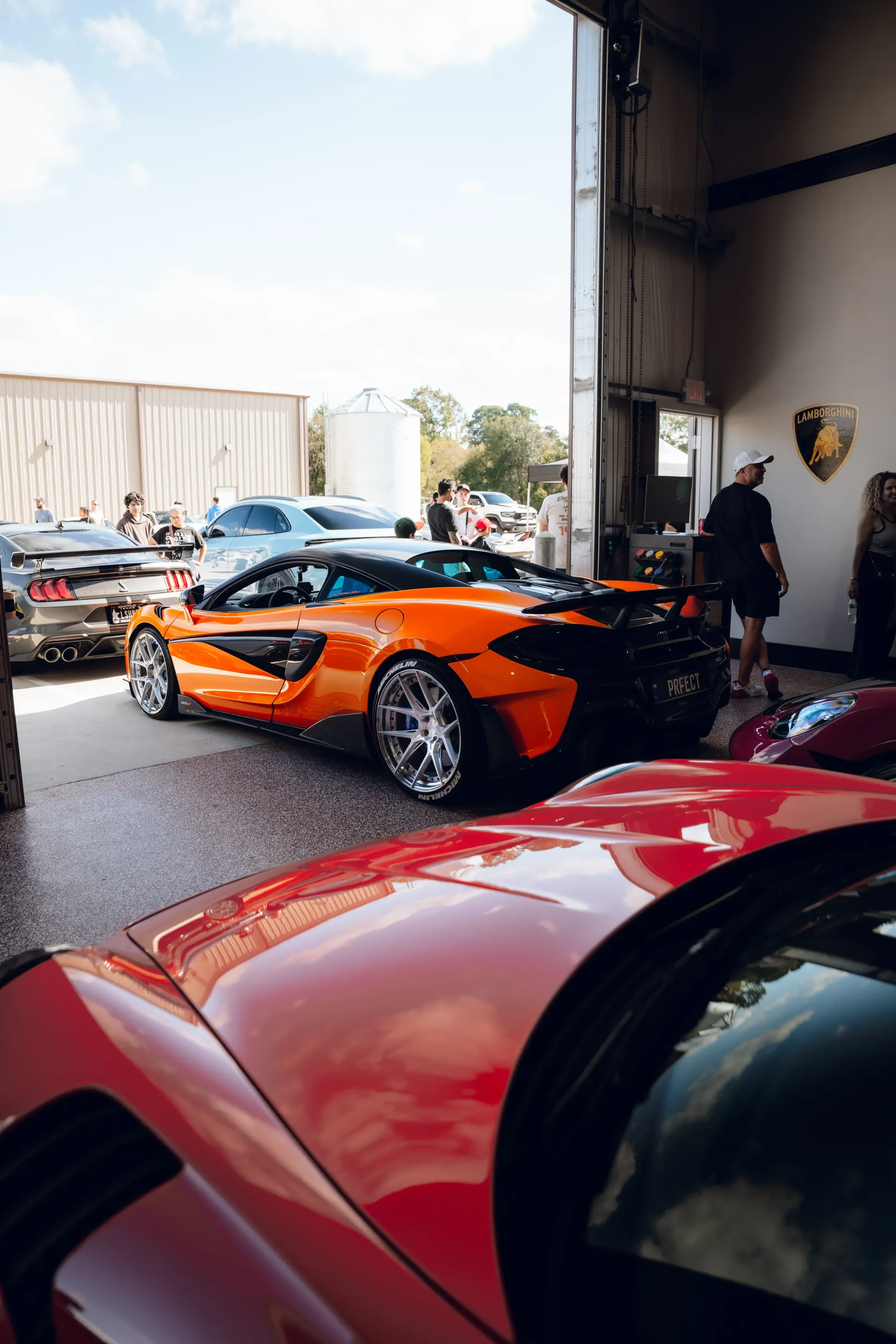 Orange McLaren sports car parked, red car in foreground, other cars and people in a garage. | Advance Automotive