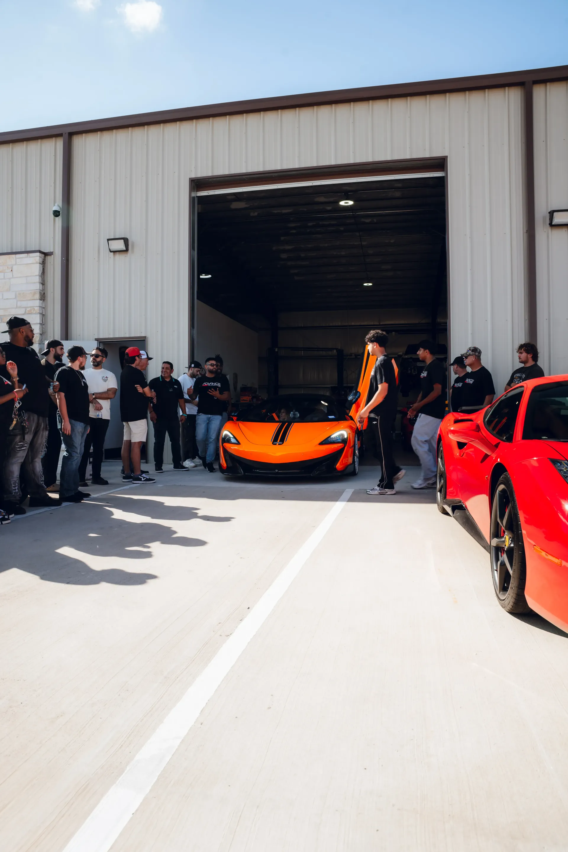 Orange sports car exits garage, flanked by onlookers and a red car. Bright sunlight. | Advance Automotive
