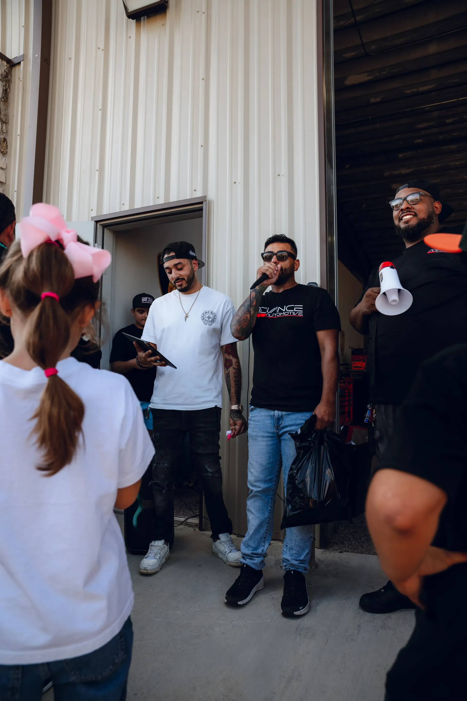 Group of people standing near a doorway. Two men in t-shirts are in the center, one holding a phone, the other a black bag. | Advance Automotive