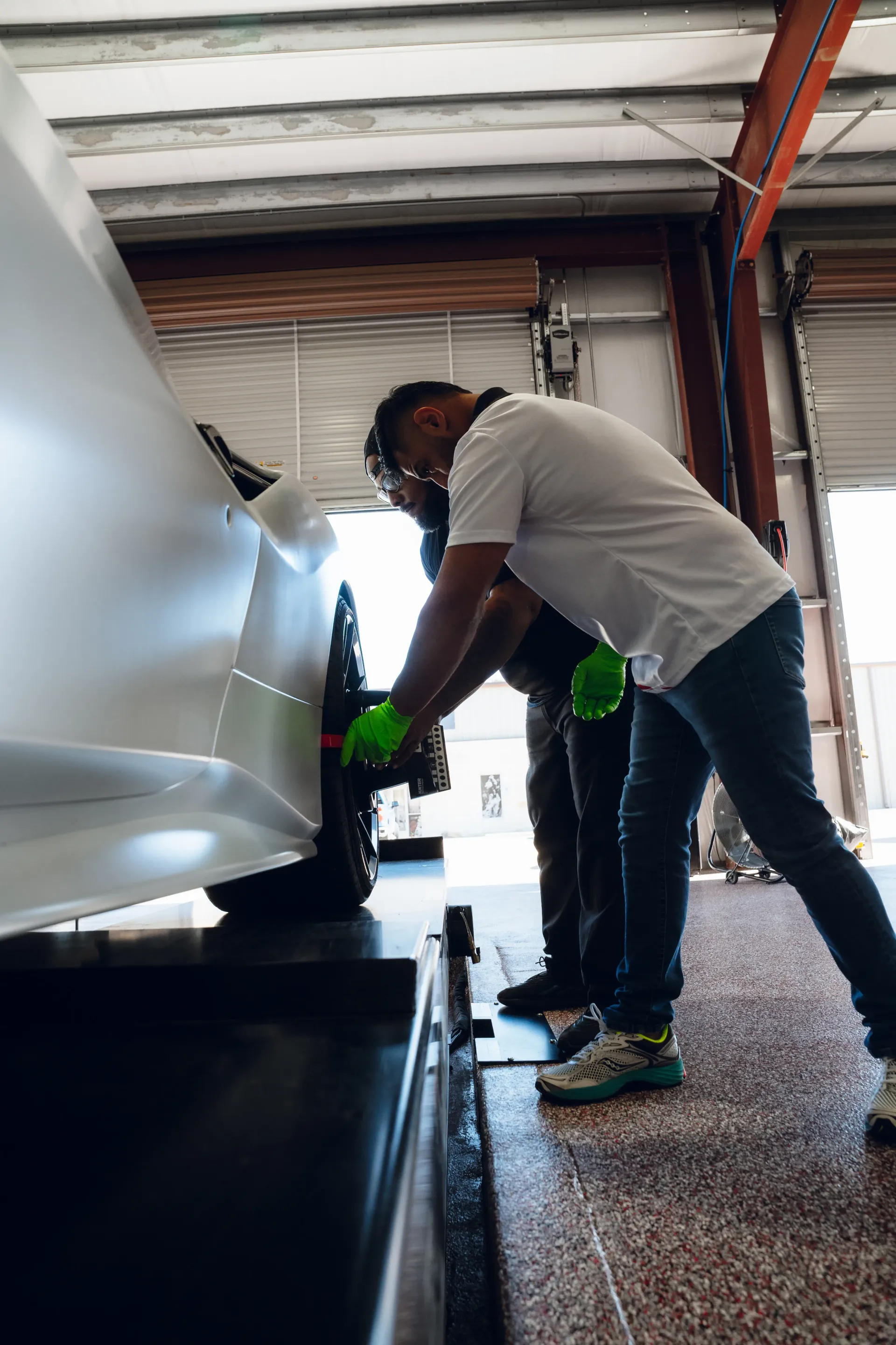 Two people working on a white car's wheel in a garage. One wears green gloves. | Advance Automotive