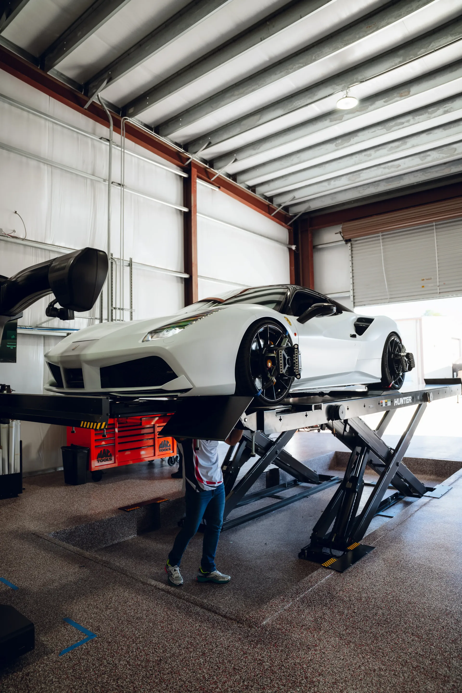 White sports car on a lift in a garage. Mechanic inspects the undercarriage. Red toolbox and workshop setting. | Advance Automotive