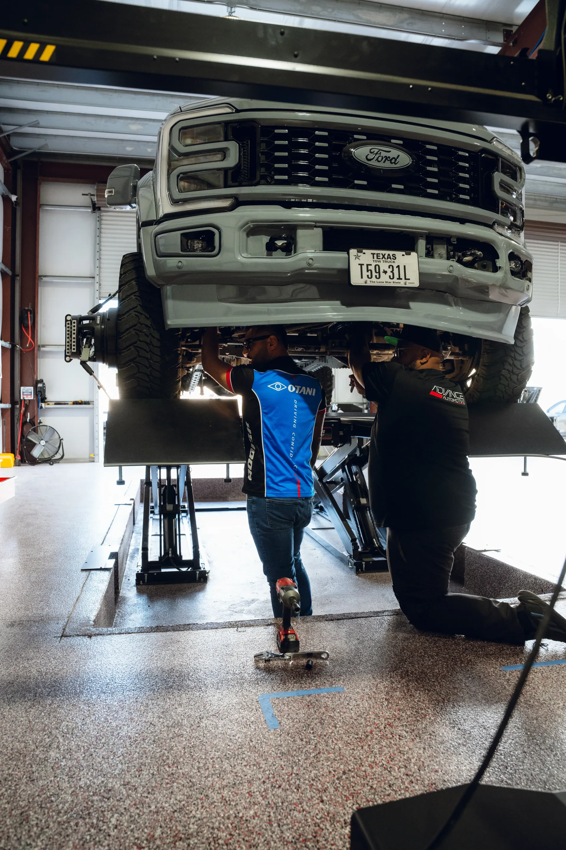 Two mechanics working on a lifted gray truck in an auto shop. The truck is suspended on a lift. | Advance Automotive