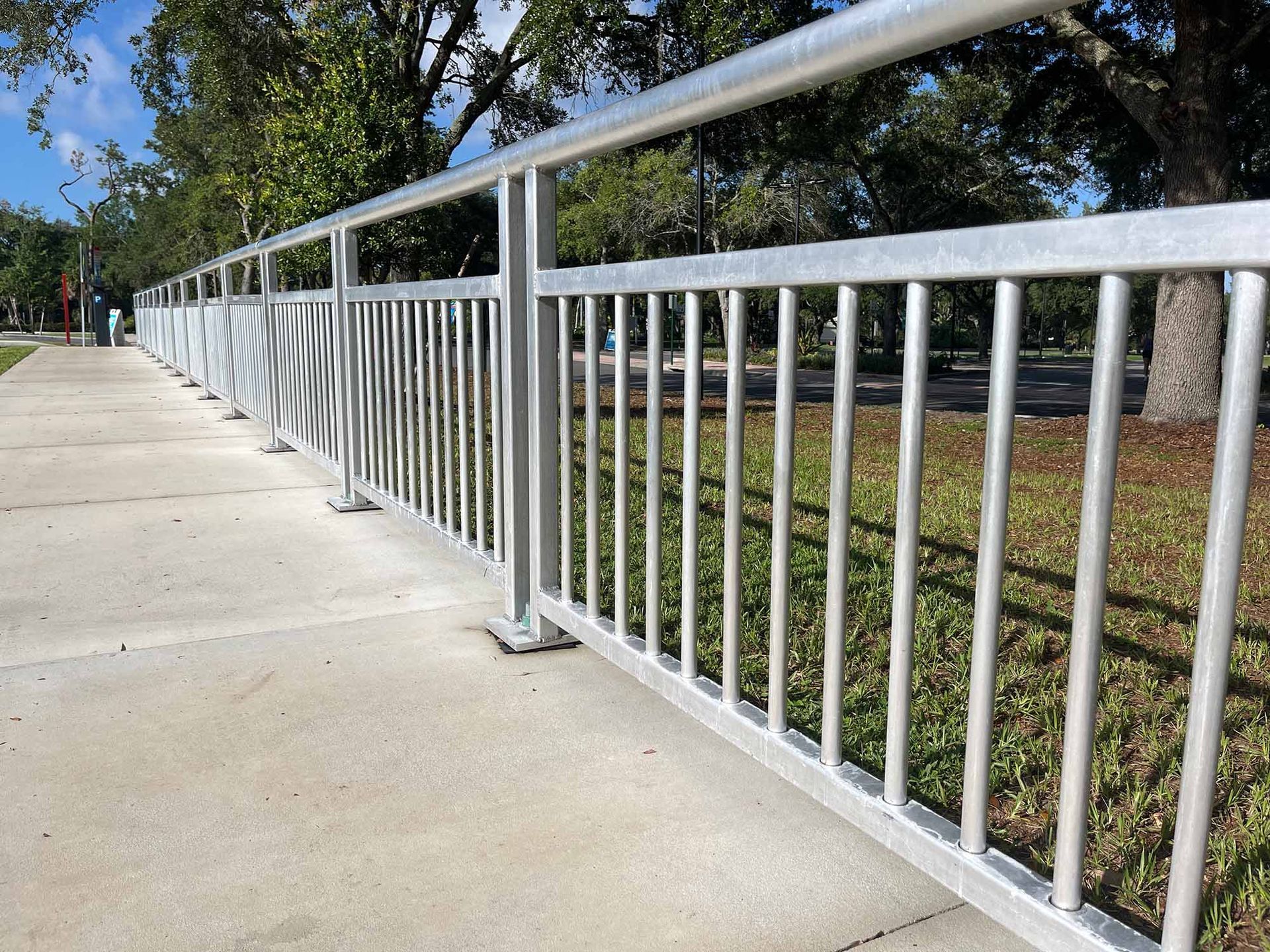 White metal railing along a sunny concrete walkway beside grass and trees