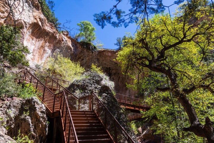 A rusty metal staircase leads up a rocky cliff face, with green trees and blue sky above.