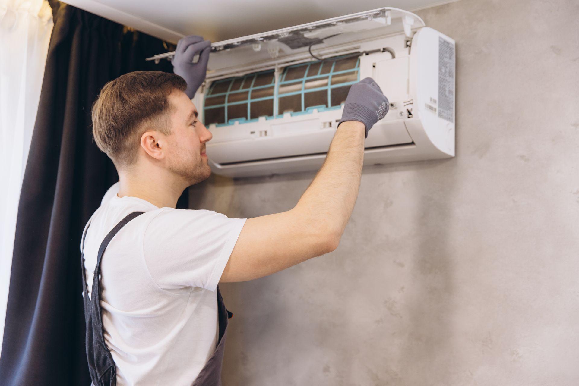 Man in gloves, fixing wall-mounted air conditioner; interior setting, gray wall. Man in gloves, fixing wall-mounted air conditioner; interior setting, gray wall.