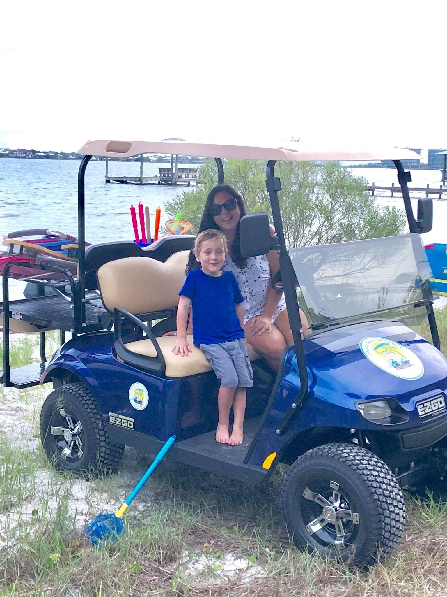 A person and a child sit on a blue golf cart parked on a grassy beach with a water view.