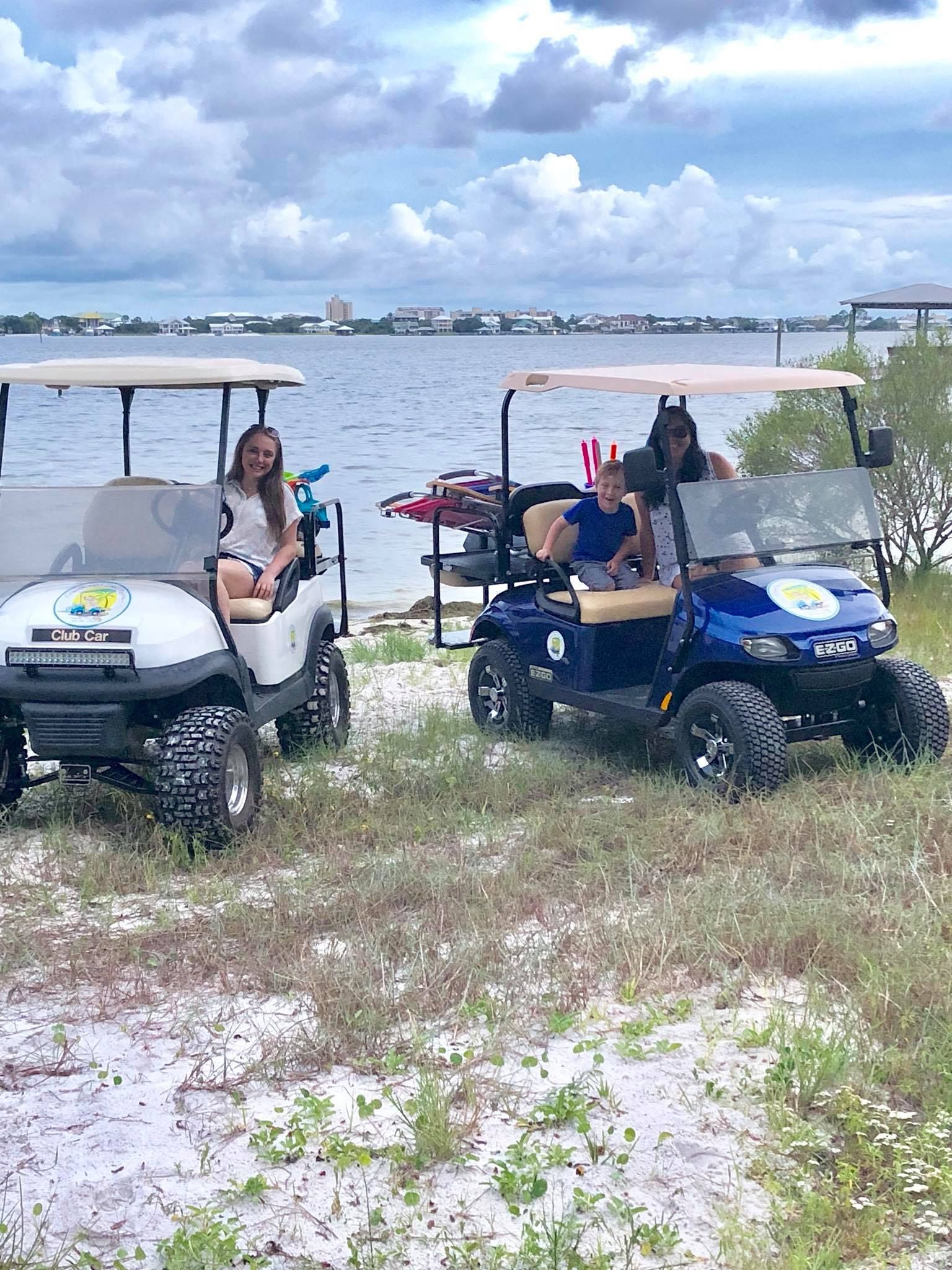 Two people sit in golf carts parked on sandy ground near a body of water under a cloudy sky.