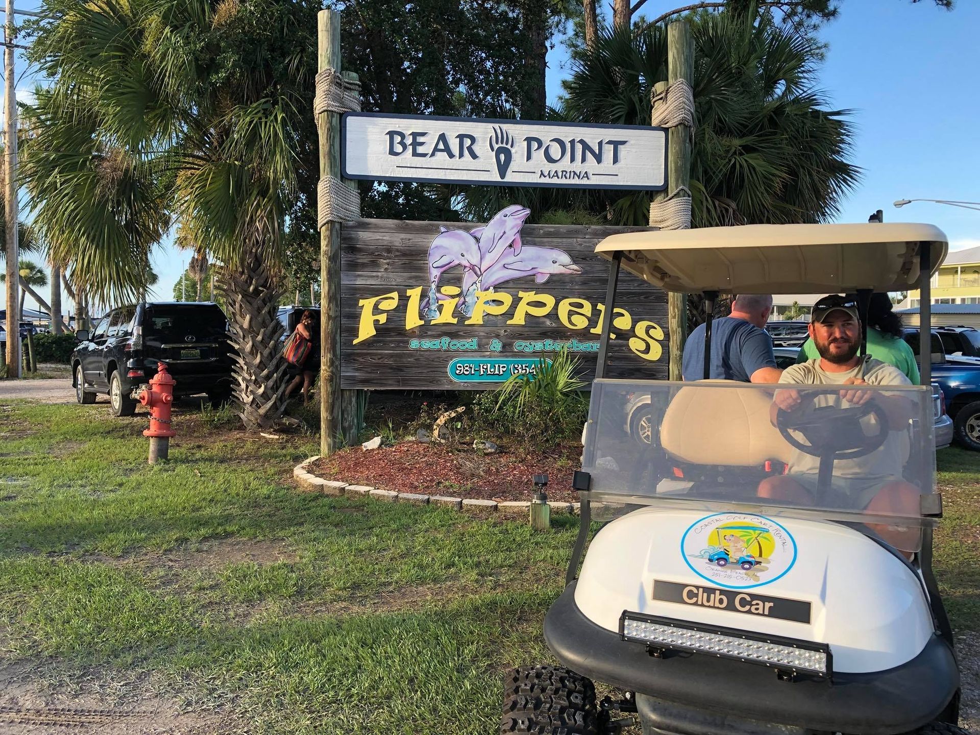 A man sits in a white golf cart in front of a wooden sign for 