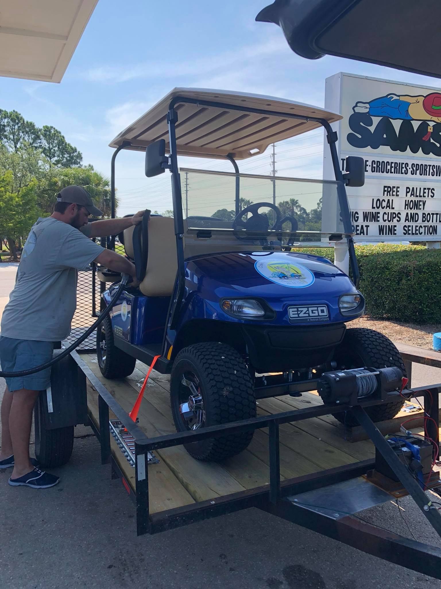 A person secures a blue golf cart to a flatbed trailer at a business parking lot on a sunny day.